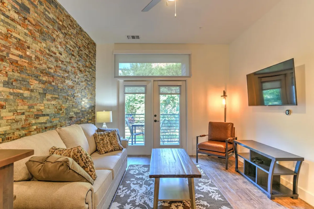 This image presents a cozy living area featuring a plush sofa, a stylish stone accent wall, a modern coffee table, and a smart TV. Ample natural light streams through the glass balcony doors, and the space is designed for relaxing, entertaining, or watching TV—matching the open-concept atmosphere described in the section.