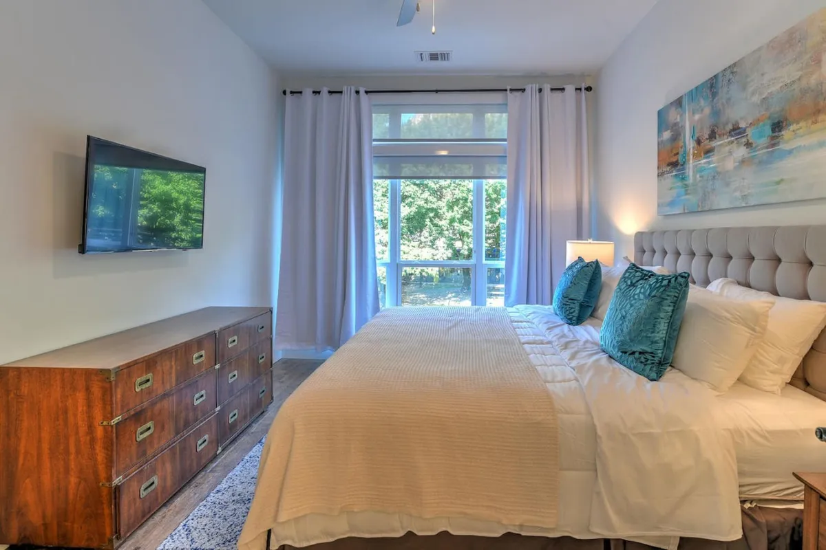 A serene bedroom with a king bed adorned with decorative pillows, a large window letting in abundant sunlight, a modern dresser for ample storage, and a television for entertainment. The décor is understated and calming, with a focus on comfort and privacy.