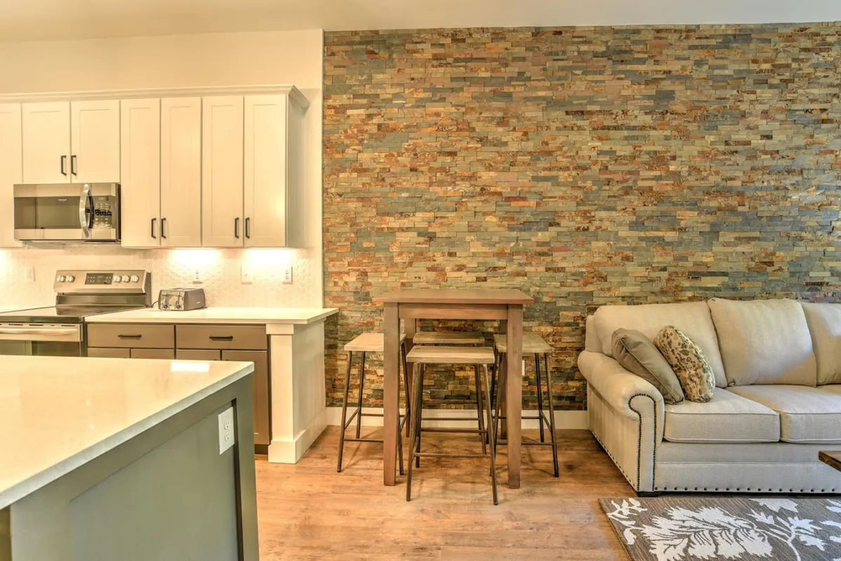 This image seamlessly captures the flow from the living area into the contemporary kitchen and dining space. The stone accent wall, dining table, and modern kitchen appliances are all visible, highlighting the open-concept layout ideal for socializing, cooking, and shared meals.