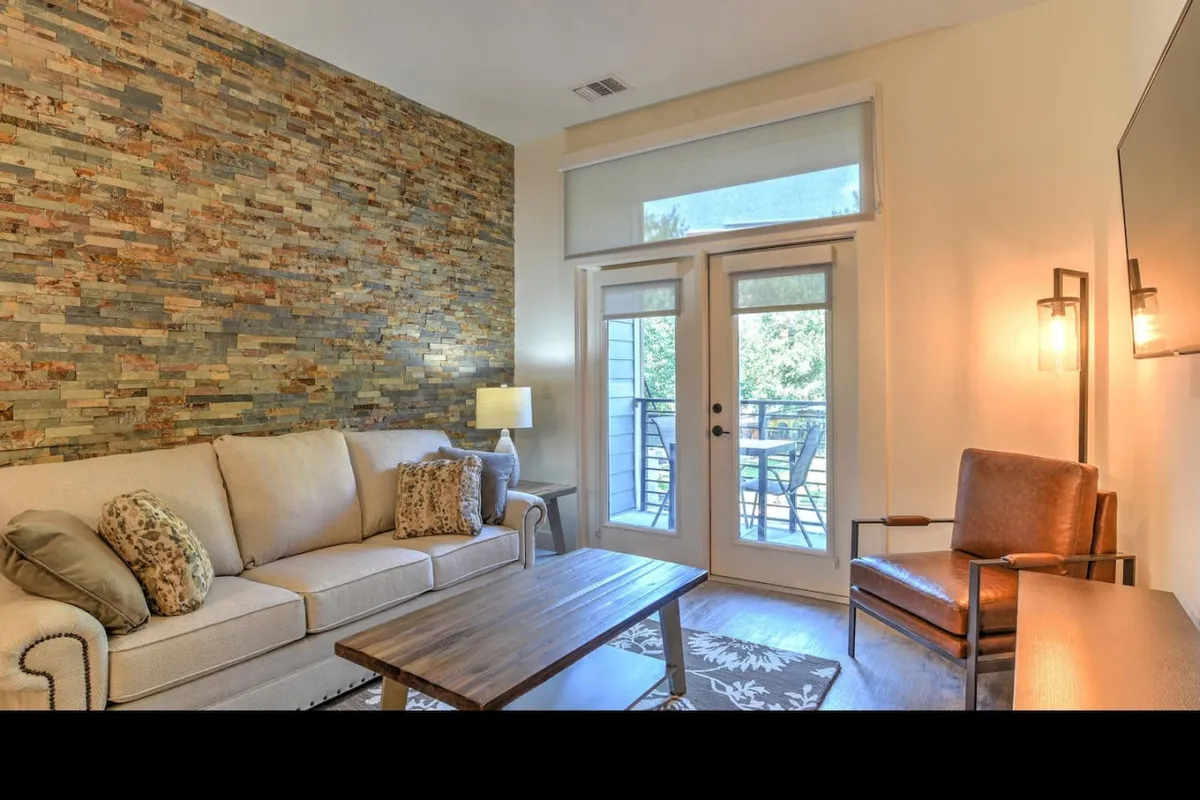 The photo looks from the interior living space out toward glass sliding doors that open to the balcony, revealing a cozy sofa, armchair, and coffee table. The arrangement draws attention to the balcony access, suggesting an easy transition from relaxing indoors to stepping outside.