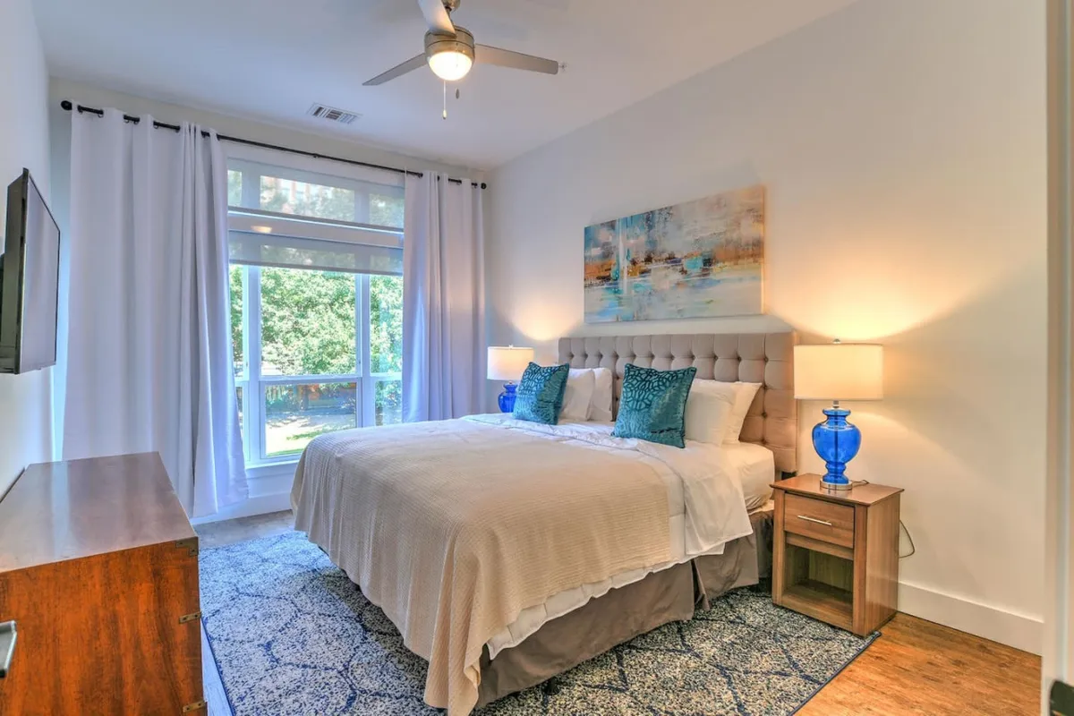 A spacious bedroom featuring a plush king-size bed dressed in crisp linens, flanked by elegant blue lamps and accented with tasteful artwork. Natural light pours in through the window, creating a calm and restful atmosphere perfect for unwinding or catching up on a good book.