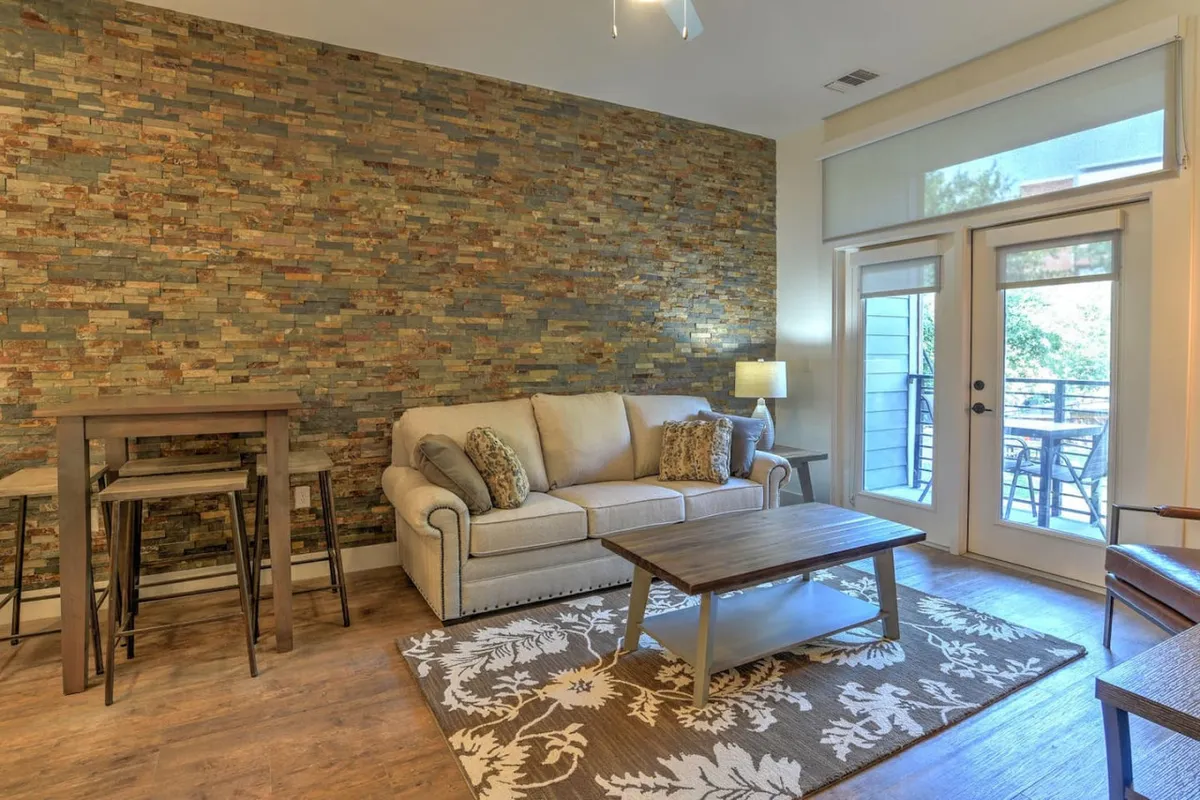 The image showcases a stylish, open-concept living area featuring a striking stone accent wall, a plush sofa adorned with decorative pillows, a warm wooden coffee table, and a vibrant floral rug. Sunlight pours in through sliding glass doors, inviting guests to step out onto the private balcony. The scene is set for cozy relaxation or socializing, with thoughtful touches that create a welcoming, homey ambiance.